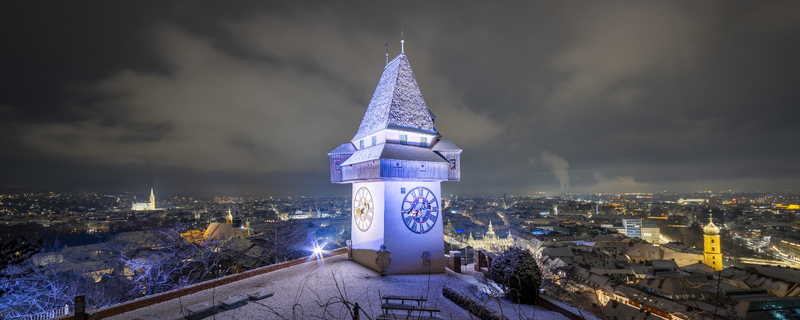 View of the iconic clock tower illuminated against the night sky standing tall over the cityscape dusted with snow a picturesque scene Graz Styria Austria. View of the iconic clock tower illuminated against the night sky standing tall over the cityscape dusted with snow a picturesque scene Graz Styria Austria.
