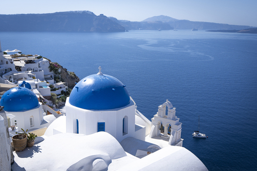 View of iconic blue domed churches overlooking the Aegean Sea with white washed buildings and a sailboat in the distance Fira Greece.