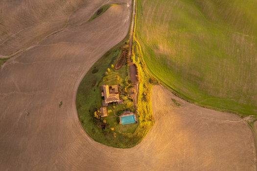 Aerial view of a traditional Tuscan farmhouse with a swimming pool surrounded by rolling hills and agricultural fields during golden hour in Asciano Tuscany Italy.