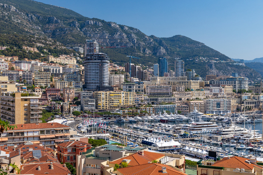 View of the harbor and cityscape of Monaco City with luxury yachts docked at Port Hercules against a backdrop of mountains and clear blue sky in Monaco City Monaco. View of the harbor and cityscape of Monaco City with luxury yachts docked at Port Hercules against a backdrop of mountains and clear blue sky in Monaco City Monaco.