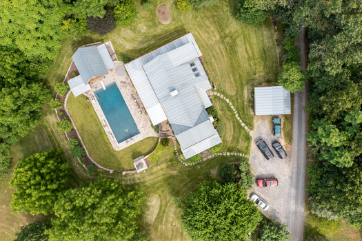 Harrisonburg Virginia United States 09 August 2021 Aerial view of a house with a metal roof a swimming pool a gravel driveway with parked cars and surrounding lush green trees. Harrisonburg Virginia United States 09 August 2021 Aerial view of a house with a metal roof a swimming pool a gravel driveway with parked cars and surrounding lush green trees.