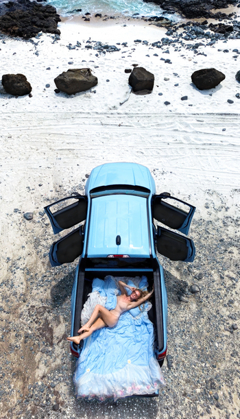 Aerial view of a woman relaxing on a bed in the back of a light blue pickup truck parked on the sandy shore of Sandy Beach in Honolulu Hawaii United States.