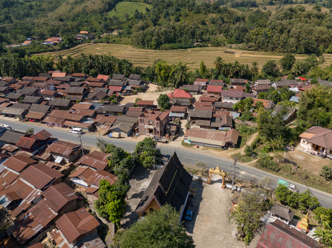 Aerial view of a village with traditional houses a temple with a dark roof and surrounding lush green hills in Ban Hat Kho Luang Prabang Province Laos.