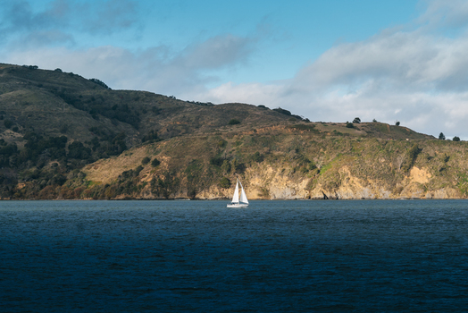 View of a white sailboat sailing on calm water with rolling coastal hills in the background under a partly cloudy sky in the United States of America. View of a white sailboat sailing on calm water with rolling coastal hills in the background under a partly cloudy sky in the United States of America.