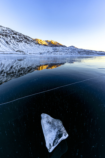 View of a piece of clear ice on a frozen lake surface with snow capped mountains reflecting in the dark water under a clear blue sky Madesimo Lombardy Italy. View of a piece of clear ice on a frozen lake surface with snow capped mountains reflecting in the dark water under a clear blue sky Madesimo Lombardy Italy.