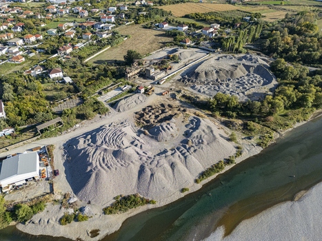 Aerial view of an industrial gravel extraction site along the riverbank with large piles of aggregate machinery and nearby residential buildings in Bahcallek Shkoder County Albania. Aerial view of an industrial gravel extraction site along the riverbank with large piles of aggregate machinery and nearby residential buildings in Bahcallek Shkoder County Albania.