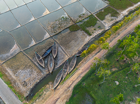 Aerial view of traditional wooden boats docked in a narrow canal beside salt pans and a tree lined path in Moheskhali Upazila Chittagong Division Bangladesh. Aerial view of traditional wooden boats docked in a narrow canal beside salt pans and a tree lined path in Moheskhali Upazila Chittagong Division Bangladesh.