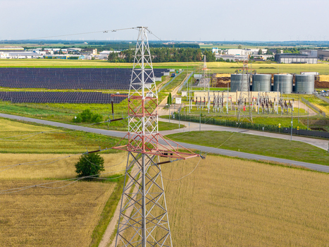 Aerial view of a high voltage electricity pylon standing amidst agricultural fields with a solar panel farm and an electrical substation in the background in Poland.