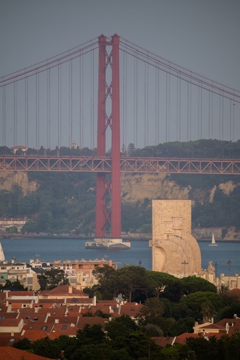 Aerial view of the Monument to the Discoveries and the 25 April Bridge spanning the Tagus River with urban rooftops and greenery in Lisboa Lisboa Portugal.
