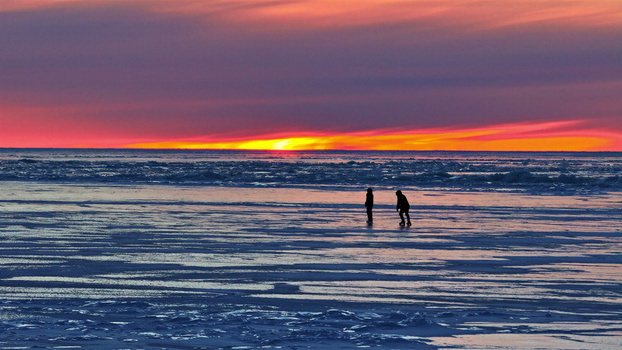 Aerial view of two people walking on a frozen icy surface during a vibrant sunset in Latvia. Aerial view of two people walking on a frozen icy surface during a vibrant sunset in Latvia.