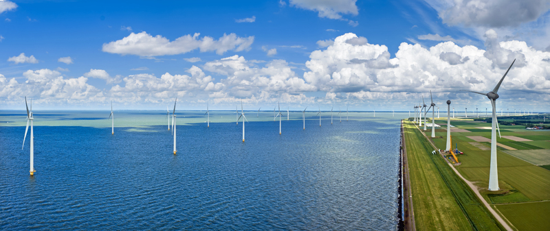 Aerial view of a large wind farm with turbines standing in the blue water and along the green fields of the coastline under a cloudy sky in Espel Flevoland Netherlands.