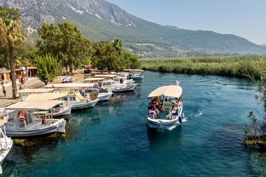 View of a boat carrying passengers on the clear blue waters of the Azmak River with docked boats along the bank lush vegetation and mountains in the background in Akyaka Mugla Turkey. View of a boat carrying passengers on the clear blue waters of the Azmak River with docked boats along the bank lush vegetation and mountains in the background in Akyaka Mugla Turkey.