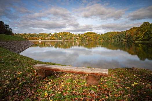 View of Velka Richnava lake with a wooden bench on the grassy shore surrounded by autumn trees under a cloudy sky Stiavnicke Bane Banska Bystrica Regio Slovakia.