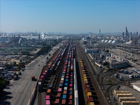 Aerial view of a busy freight railyard with colorful shipping containers on trains adjacent to an industrial complex with storage tanks in Carson California United States.