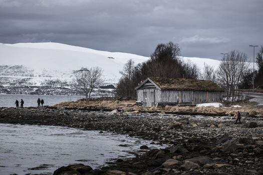 View of a traditional wooden boathouse with a sod roof on a rocky shoreline with people walking near the water and snow capped mountains in the distance Tromso Troms Norway.