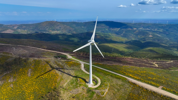 Aerial view of a wind turbine standing on a grassy hill with yellow wildflowers surrounded by rolling hills and a dirt road in Lisbon Lisbon Portugal. Aerial view of a wind turbine standing on a grassy hill with yellow wildflowers surrounded by rolling hills and a dirt road in Lisbon Lisbon Portugal.