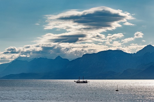 View of a small boat sailing on the calm sea with the majestic Taurus Mountains in the background under a dramatic cloudy sky in Antalya Antalya Turkey. View of a small boat sailing on the calm sea with the majestic Taurus Mountains in the background under a dramatic cloudy sky in Antalya Antalya Turkey.
