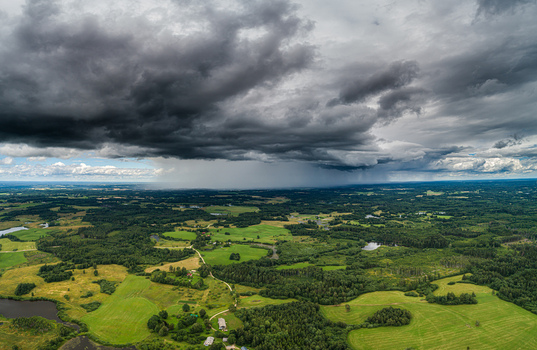 Aerial view of a dramatic dark storm cloud hanging over a vast green landscape featuring lush forests agricultural fields and small ponds in Latvia. Aerial view of a dramatic dark storm cloud hanging over a vast green landscape featuring lush forests agricultural fields and small ponds in Latvia.