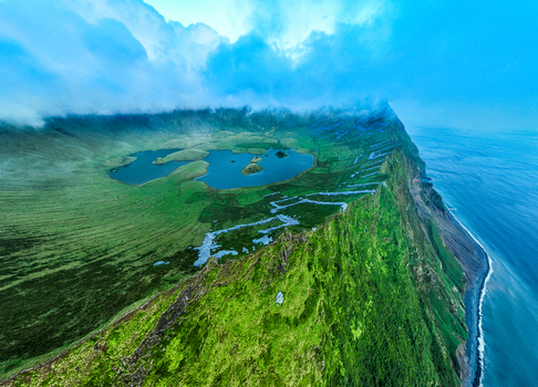 Aerial view of the volcanic crater lake of Caldeirao on Corvo Island surrounded by lush green slopes rugged cliffs and the Atlantic Ocean in Vila do Corvo Azores Portugal.
