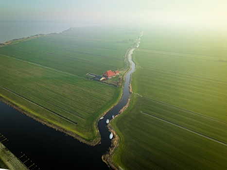 Aerial view of a narrow winding canal through green polders with a farmhouse and small boats under a hazy sky Sloten Friesland Netherlands. Aerial view of a narrow winding canal through green polders with a farmhouse and small boats under a hazy sky Sloten Friesland Netherlands.