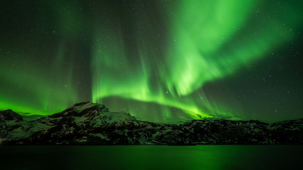 View of the vibrant green Aurora Borealis illuminating the night sky above the rugged snow capped mountains and dark waters in Leknes Lofoten Norway.