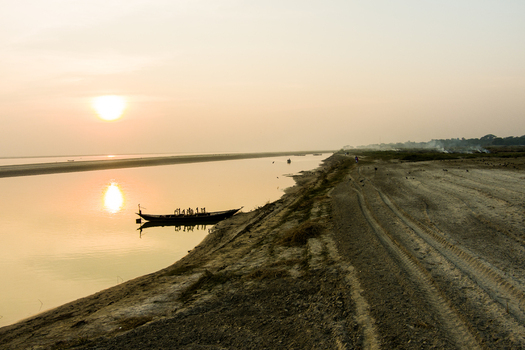 Aerial view of a traditional wooden boat floating on the calm Padma River during a golden sunset with tire tracks visible on the sandy riverbank in Rajshahi Rajshahi Division Bangladesh.