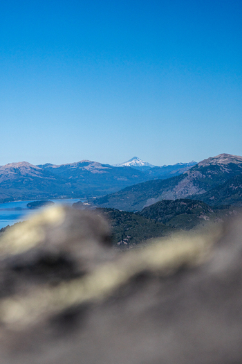 View of the snow capped Lanin volcano rising above mountains and a blue lake with a blurred rocky foreground in Huiliches Department Neuquen Province Argentina. View of the snow capped Lanin volcano rising above mountains and a blue lake with a blurred rocky foreground in Huiliches Department Neuquen Province Argentina.