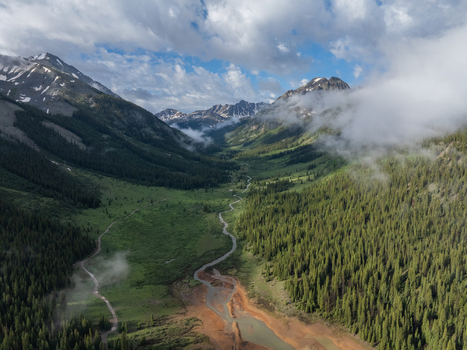 View of a winding river flowing through a lush green mountain valley surrounded by dense pine forests and snow capped peaks under a cloudy sky in Colorado United States.