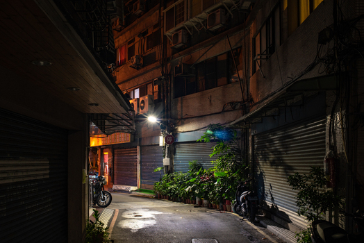 View of a narrow alleyway in Ximen at night featuring closed metal shutters of shops potted plants a parked scooter and overhead air conditioning units in Ximen Taipei City Taiwan. View of a narrow alleyway in Ximen at night featuring closed metal shutters of shops potted plants a parked scooter and overhead air conditioning units in Ximen Taipei City Taiwan.