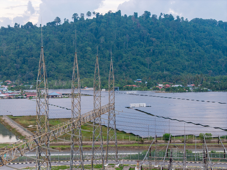 Aerial view of a high voltage transmission substation with metal towers and a large solar panel farm set against a lush green hill in Central Seberang Perai Penang Malaysia. Aerial view of a high voltage transmission substation with metal towers and a large solar panel farm set against a lush green hill in Central Seberang Perai Penang Malaysia.