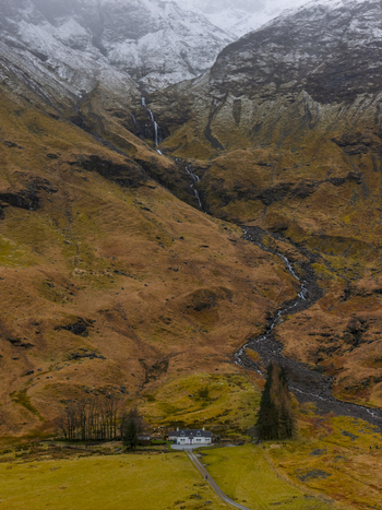 Aerial view of a lonely white cottage at the base of rugged mountains with a cascading waterfall and snow capped peaks Glencoe Scotland United Kingdom. Aerial view of a lonely white cottage at the base of rugged mountains with a cascading waterfall and snow capped peaks Glencoe Scotland United Kingdom.