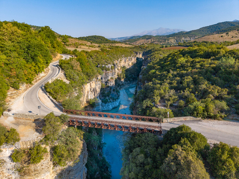 Aerial view of Osumi Canyon featuring a metal truss bridge spanning the turquoise river surrounded by winding roads and lush green hills in Dhores Berat County Albania.