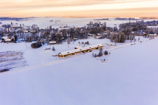 Aerial view of a rural settlement with buildings covered in snow surrounded by vast frozen fields and misty forests under a soft golden sunset sky in Latvia. Aerial view of a rural settlement with buildings covered in snow surrounded by vast frozen fields and misty forests under a soft golden sunset sky in Latvia.