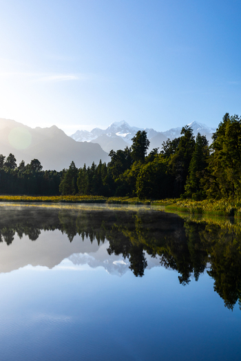 View of mirror like lake reflecting the silhouetted forest and distant snow capped mountains under a clear sky Lake Matheson New Zealand. View of mirror like lake reflecting the silhouetted forest and distant snow capped mountains under a clear sky Lake Matheson New Zealand.