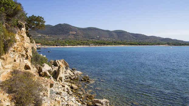 View of rugged cliffs meet the tranquil turquoise sea under a clear blue sky as lush green hills rise in the distance Lacona Elba Italy. View of rugged cliffs meet the tranquil turquoise sea under a clear blue sky as lush green hills rise in the distance Lacona Elba Italy.
