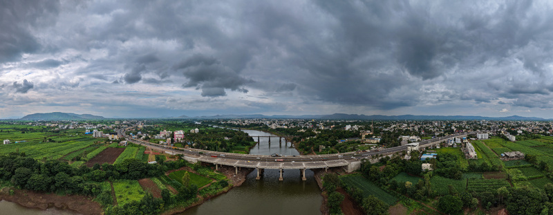Aerial view of the bridge spanning the river in Karad surrounded by lush green agricultural fields and urban development under a dramatic cloudy sky in Karad Maharashtra India. Aerial view of the bridge spanning the river in Karad surrounded by lush green agricultural fields and urban development under a dramatic cloudy sky in Karad Maharashtra India.