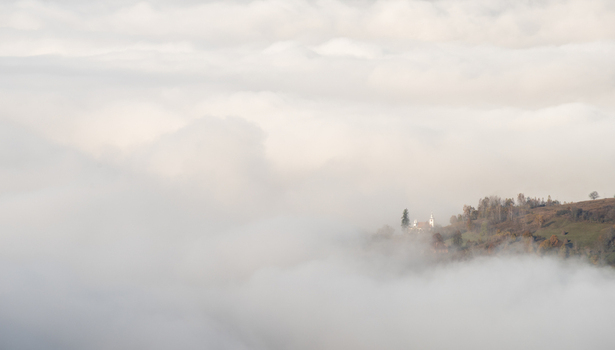 View of a church perched on a hillside emerging from a thick layer of morning fog with autumn trees and soft light in Alba Iulia Alba County Romania. View of a church perched on a hillside emerging from a thick layer of morning fog with autumn trees and soft light in Alba Iulia Alba County Romania.