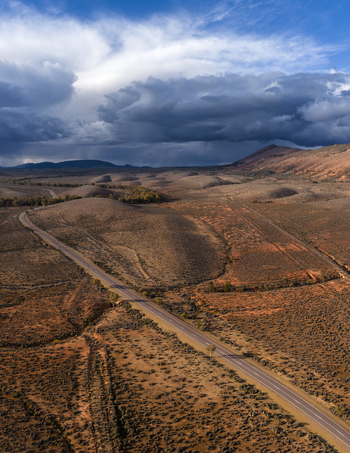 Aerial view of a long asphalt road winding through the arid outback landscape under dramatic storm clouds and rolling hills in Flinders Ranges South Australia Australia.
