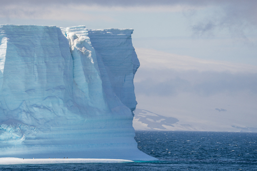 View of a massive blue iceberg floating in the Southern Ocean near Paulet Island with distant snow covered mountains and small penguins standing on the ice base Antarctica. View of a massive blue iceberg floating in the Southern Ocean near Paulet Island with distant snow covered mountains and small penguins standing on the ice base Antarctica.