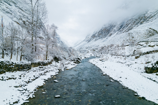 Aerial view of a flowing river winding through a snow covered valley surrounded by rocky mountain slopes and sparse trees in Nepal.
