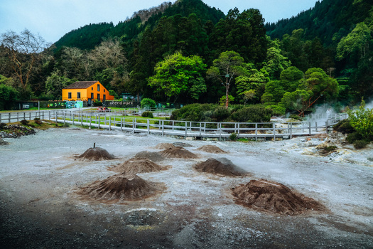 View of steaming fumaroles dotting the volcanic landscape juxtaposed against a vibrant yellow building nestled among lush green trees Furnas Azores Portugal.
