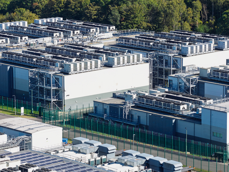 Aerial view of a large data center facility with rows of cooling units on the rooftops secure perimeter fencing and surrounding greenery in the United Kingdom.