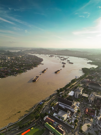 Aerial view of the Mahakam River with coal barges navigating the waterway surrounded by urban development and lush greenery in Samarinda Kalimantan Timur Indonesia. Aerial view of the Mahakam River with coal barges navigating the waterway surrounded by urban development and lush greenery in Samarinda Kalimantan Timur Indonesia.