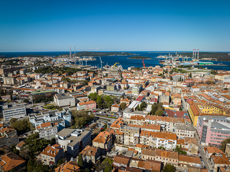 Aerial view of terracotta rooftops cascading towards the shimmering Adriatic Sea where industrial structures meet the horizon Pula Istria County Croatia.