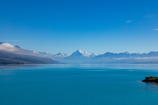 View of a turquoise lake reflecting the snow capped mountains under a clear blue sky with a small green islet accenting the tranquil scene Lake Pukaki Mount Cook Lookout New Zealand. View of a turquoise lake reflecting the snow capped mountains under a clear blue sky with a small green islet accenting the tranquil scene Lake Pukaki Mount Cook Lookout New Zealand.