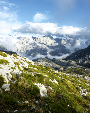 View of rugged verdant slopes lead to majestic snow dusted peaks piercing through swirling clouds and a vibrant blue sky Picos de Europa Asturias Spain. View of rugged verdant slopes lead to majestic snow dusted peaks piercing through swirling clouds and a vibrant blue sky Picos de Europa Asturias Spain.