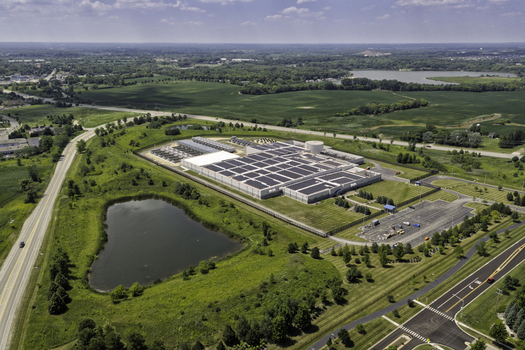 Aerial view of a large industrial facility with solar panels on the roof surrounded by green fields a small pond and nearby roads in Volo Illinois United States. Aerial view of a large industrial facility with solar panels on the roof surrounded by green fields a small pond and nearby roads in Volo Illinois United States.