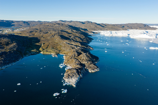 Aerial view of Ilulissat glacier with floating icebergs in the deep blue water and rugged rocky coastline under a clear sky in Greenland. Aerial view of Ilulissat glacier with floating icebergs in the deep blue water and rugged rocky coastline under a clear sky in Greenland.