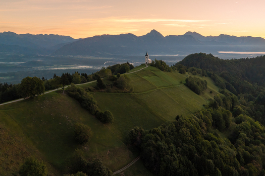 Aerial view of the Church of Saints Primus and Felician perched on a grassy hill with a winding path and mountain backdrop during golden hour in Jamnik Kranj Slovenia.