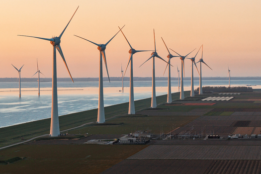 Aerial view of wind turbines standing in a row along a dike between agricultural fields and the sea during a golden sunset Netherlands.